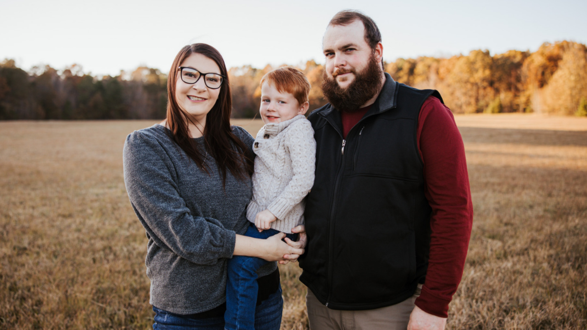 A woman with glasses and a man with a beard stand in a golden field at sunset, smiling at the camera. The woman holds a young boy with red hair between them.